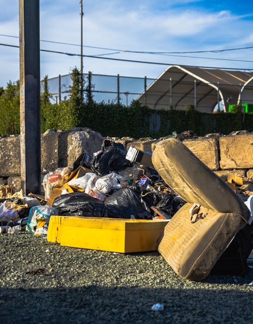 An outdoor scene showing a pile of mixed rubbish and discarded items on a gravel surface, including black trash bags, a yellow plastic container, an old, dirty car tyre leaning against the rubbish, an