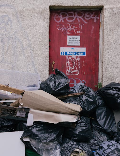 An outdoor scene showing a pile of mixed rubbish and discarded items on a gravel surface, including black trash bags, a yellow plastic container, an old, dirty car tyre leaning against the rubbish, an
